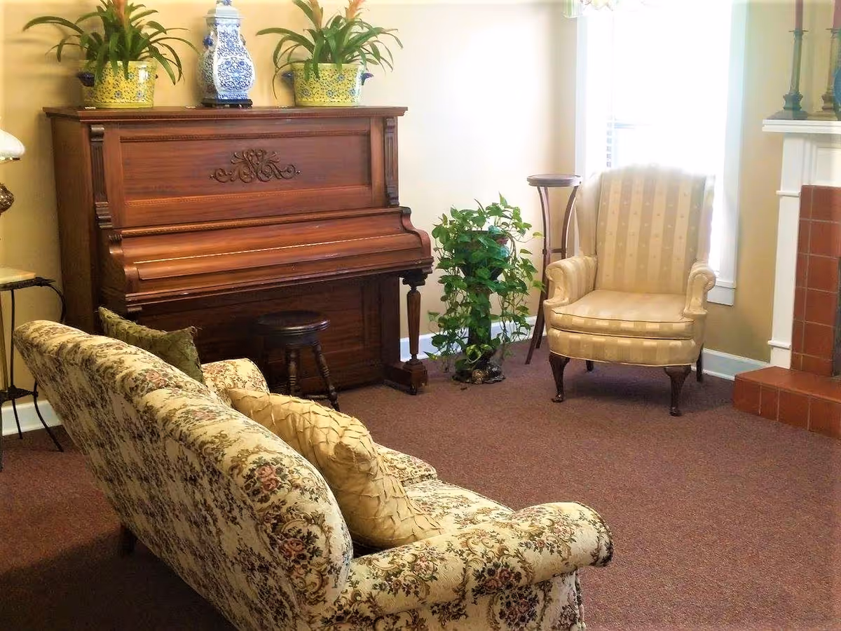 A cozy living room with a vintage upright piano adorned with two yellow floral pots and a blue and white decorative vase on top. In front of the piano is a wooden piano stool. To the left is a floral patterned sofa with two cushions, and to the right is a beige striped armchair near a window. A green potted plant sits on the floor between the piano and the armchair. The room has a carpeted floor and a fireplace with red tiles on the right side.