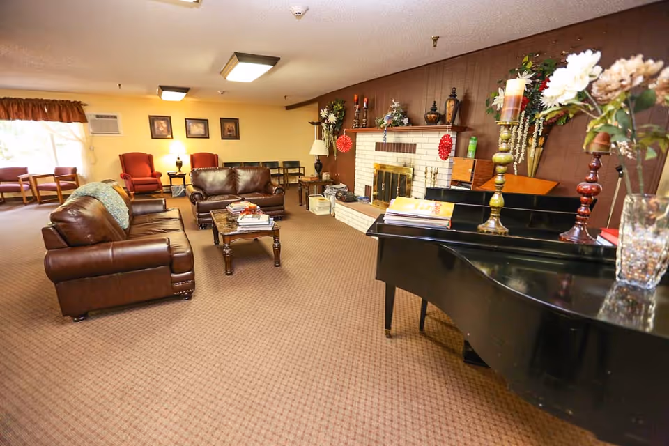 A cozy living room area with brown leather sofas, a wooden coffee table with books, a black grand piano adorned with candles and flowers, and a white brick fireplace decorated with various ornaments. The room has carpeted flooring, framed pictures on the wall, and several chairs near a window with curtains.