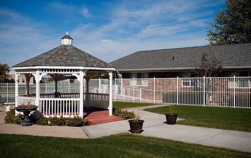 White wooden gazebo on a lawn with potted plants and a fenced single-story building in the background.