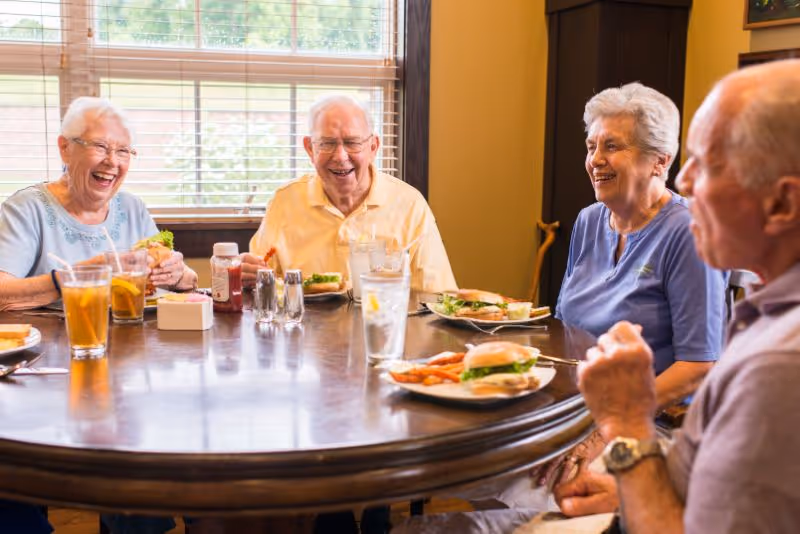 Four elderly people sitting around a round wooden dining table enjoying a meal together. The table has plates with sandwiches and fries, glasses of iced tea and water, and condiments. They are smiling and appear to be having a pleasant conversation in a well-lit room with a window in the background.