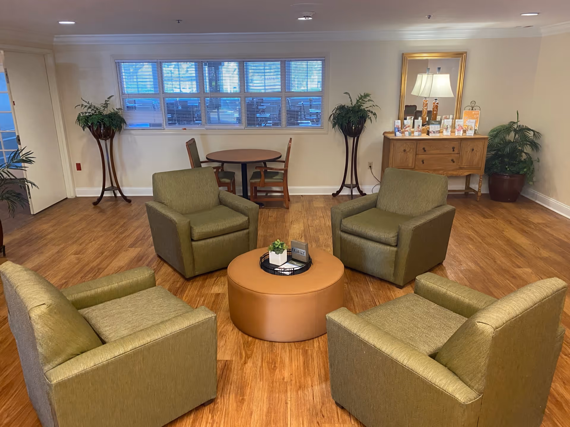 A cozy seating area in a senior living facility with four green armchairs arranged around a round brown ottoman. Behind the seating area, there is a small round table with two chairs, two tall plant stands with greenery, and a wooden sideboard with a lamp, mirror, and various brochures or pamphlets. The room has wooden flooring and a window with blinds.