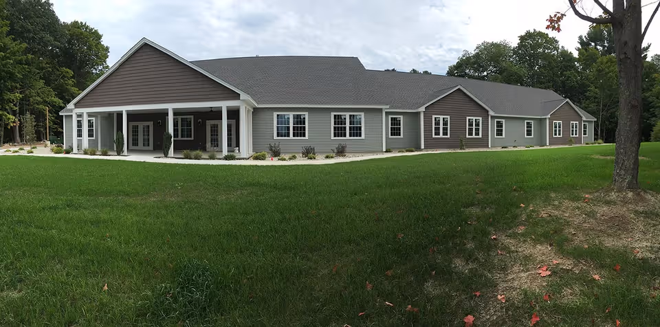 Exterior view of a single-story assisted living facility building with gray siding and white trim, surrounded by a green lawn and trees under a cloudy sky.