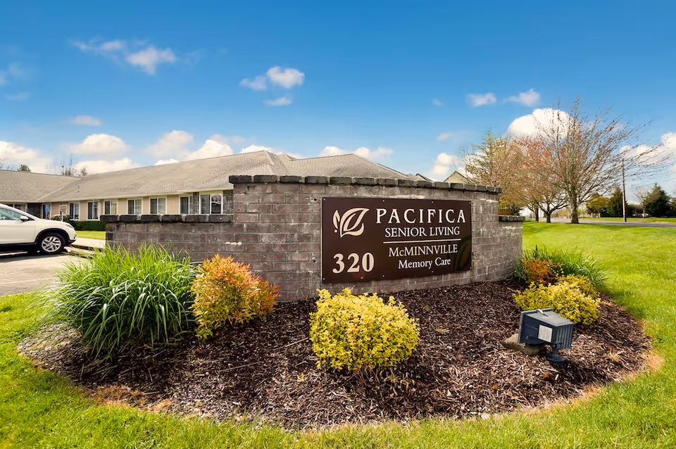 Brick entrance sign that reads 'Pacifica Senior Living McMinnville Memory Care 320' set in landscaped mulch and grass with the facility building and a parked car behind it.