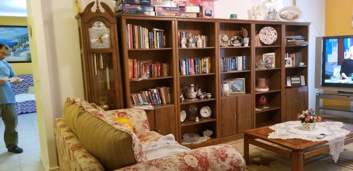A cozy living room with a floral patterned couch, a wooden coffee table with a lace doily and a small flower arrangement, a large wooden bookshelf filled with books, decorative plates, and figurines, a grandfather clock, and a television showing a person on screen. A person in a blue uniform is seen in the adjacent room.
