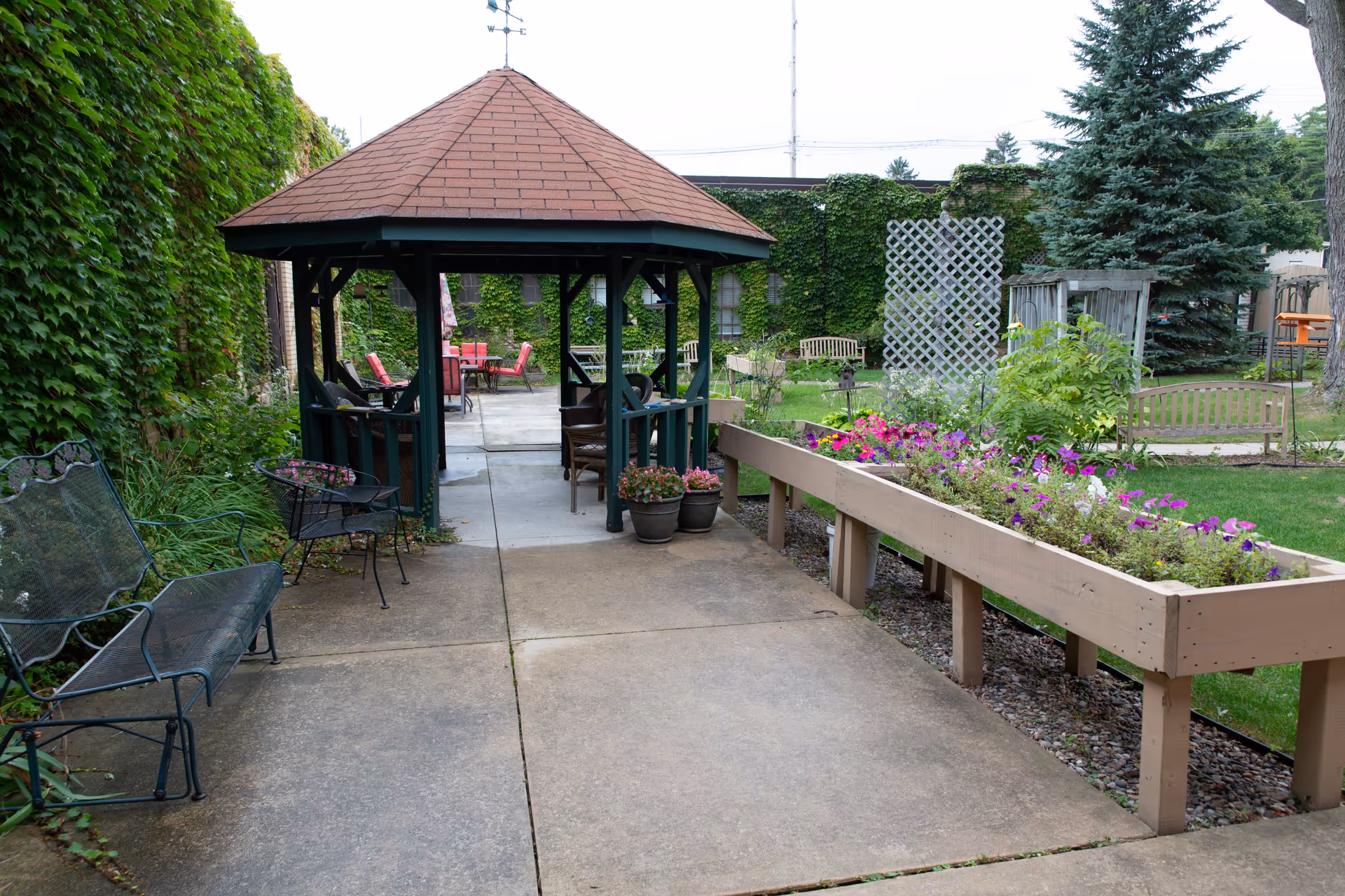 Outdoor courtyard with a wooden gazebo, benches, potted plants and raised flower beds beside a paved walkway.