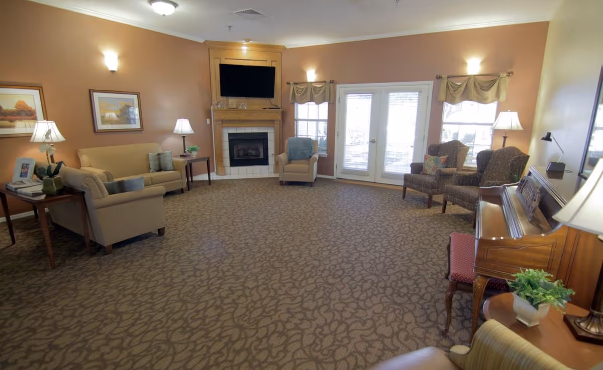 A cozy living room with beige walls and patterned carpet. The room features a beige sofa, armchairs, a wooden piano with a red cushioned bench, and a fireplace with a mounted TV above it. There are two framed pictures on the wall, several table lamps, and French doors with windows on either side letting in natural light.