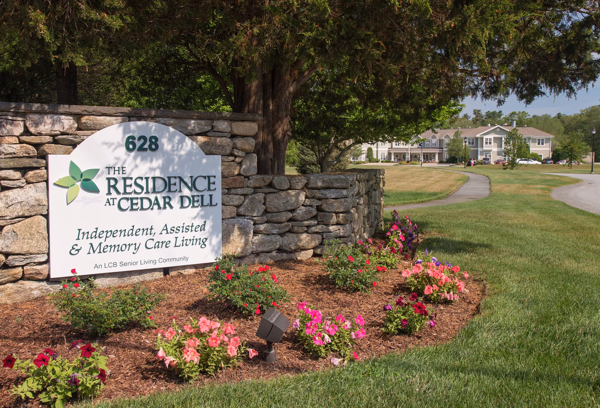 Stone sign for The Residence at Cedar Dell with flowers planted in front, a large tree behind the sign, and a senior living facility building visible in the background with a curved driveway and green lawn.