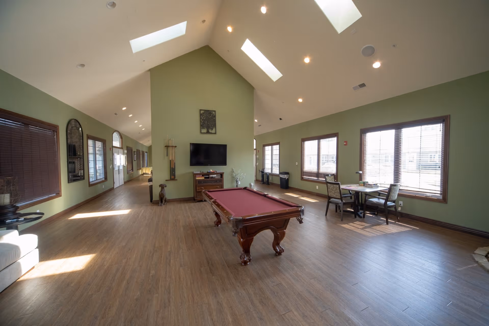 A spacious common area with green walls and wooden flooring featuring a pool table with a red felt top in the center. The room has large windows with blinds allowing natural light to fill the space. There is a wall-mounted TV above a wooden cabinet, a small table with chairs near the windows, and a few decorative items including a wall clock and a small statue of a dog. The ceiling is vaulted with skylights and recessed lighting.