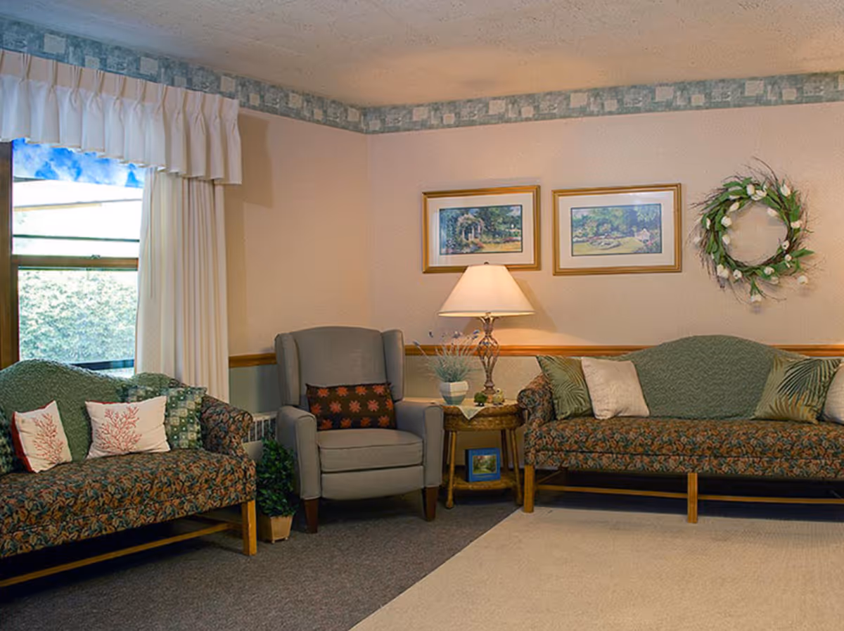 A cozy living room area with two patterned sofas and a gray armchair. The sofas have decorative pillows, and there is a wooden side table with a lamp and a small plant. Two framed landscape paintings and a floral wreath hang on the beige wall. A window with white curtains lets in natural light.