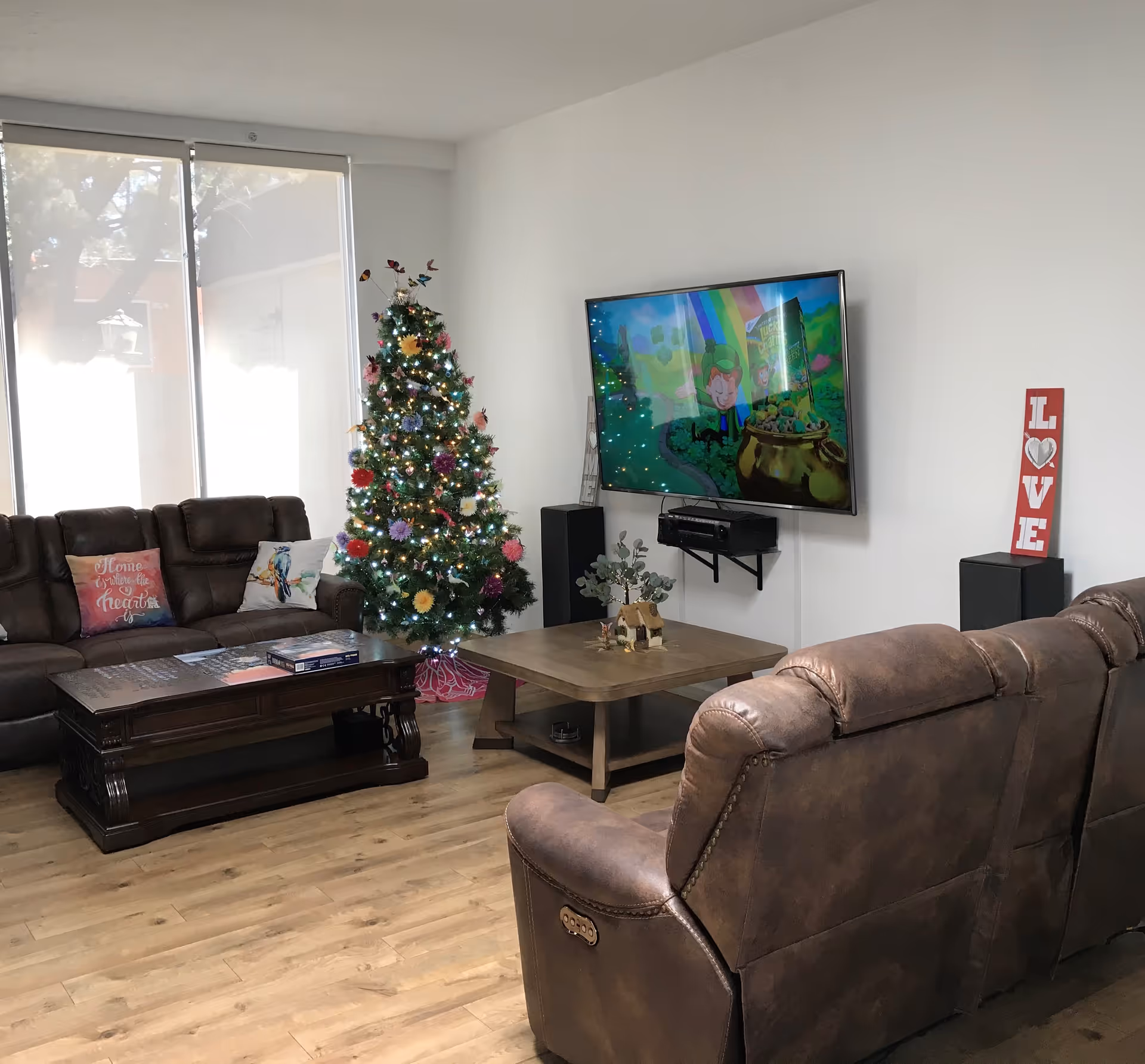 A cozy living room with brown leather sofas, a decorated Christmas tree with lights, a wooden coffee table with a small decorative house and plant, a wall-mounted flat-screen TV displaying a colorful animated scene, and a red decorative sign with the word 'LOVE' on a speaker.