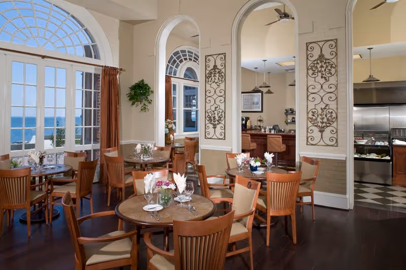 A bright dining room with round wooden tables and chairs, each table set with glasses, plates, and floral centerpieces. Large arched windows with brown curtains offer a view of the ocean outside. Decorative wall panels and an open kitchen area are visible in the background.