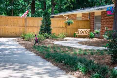 Sunlit courtyard with paved walkways, a wooden bench by a brick building, landscaped planting beds, and a wooden fence.