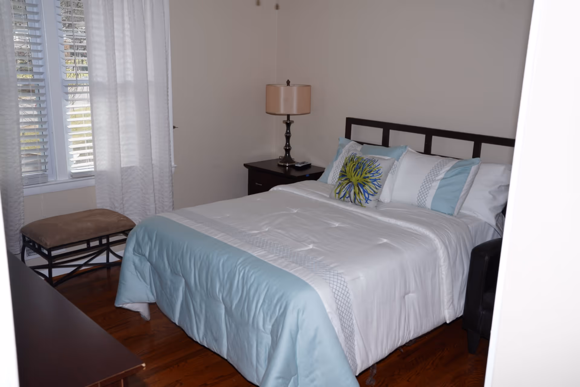 A bedroom with a neatly made bed featuring white and light blue bedding, decorative pillows, a bedside table with a lamp, a cushioned bench near a window with white curtains, and wooden flooring.