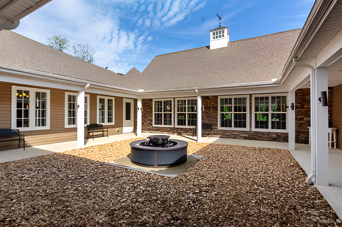 Outdoor courtyard area of a senior living facility with a central circular water fountain surrounded by gravel and concrete walkways. The courtyard is enclosed by a building with beige siding, white trim, multiple windows, and a shingled roof under a blue sky with some clouds. There are benches along the walls under a covered walkway.