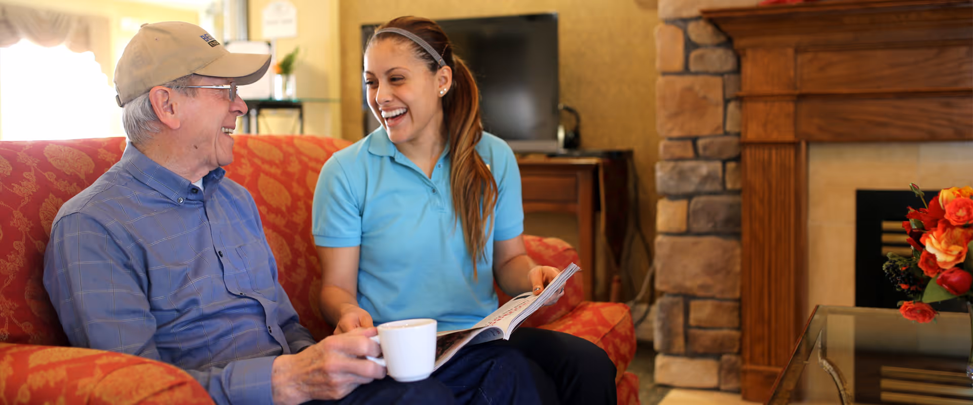 An elderly man wearing a cap and glasses sits on a red patterned couch holding a white mug, smiling and talking with a young woman in a blue polo shirt who is holding a magazine. They are in a cozy living room with a stone fireplace, wooden mantel, and a vase of red and orange flowers on a glass table.
