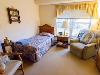 A cozy bedroom with a single bed covered in a floral quilt, a wooden headboard, and a folded towel on top. Next to the bed is a small wooden nightstand with a lamp, clock, and other small items. A large window with yellow curtains lets in natural light. There is a comfortable armchair with a patterned throw blanket and a wooden chair with a cushion. The room has beige carpet and light-colored walls.