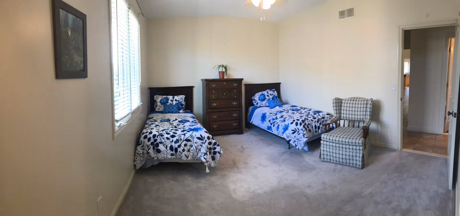 A bedroom with two single beds featuring blue and white floral bedding. Between the beds is a wooden chest of drawers with a small potted plant on top. To the right of the beds is a cushioned armchair with a plaid pattern. The room has beige walls, a window with blinds on the left wall, and a ceiling fan with lights. There is an open doorway leading to another room with tiled flooring.