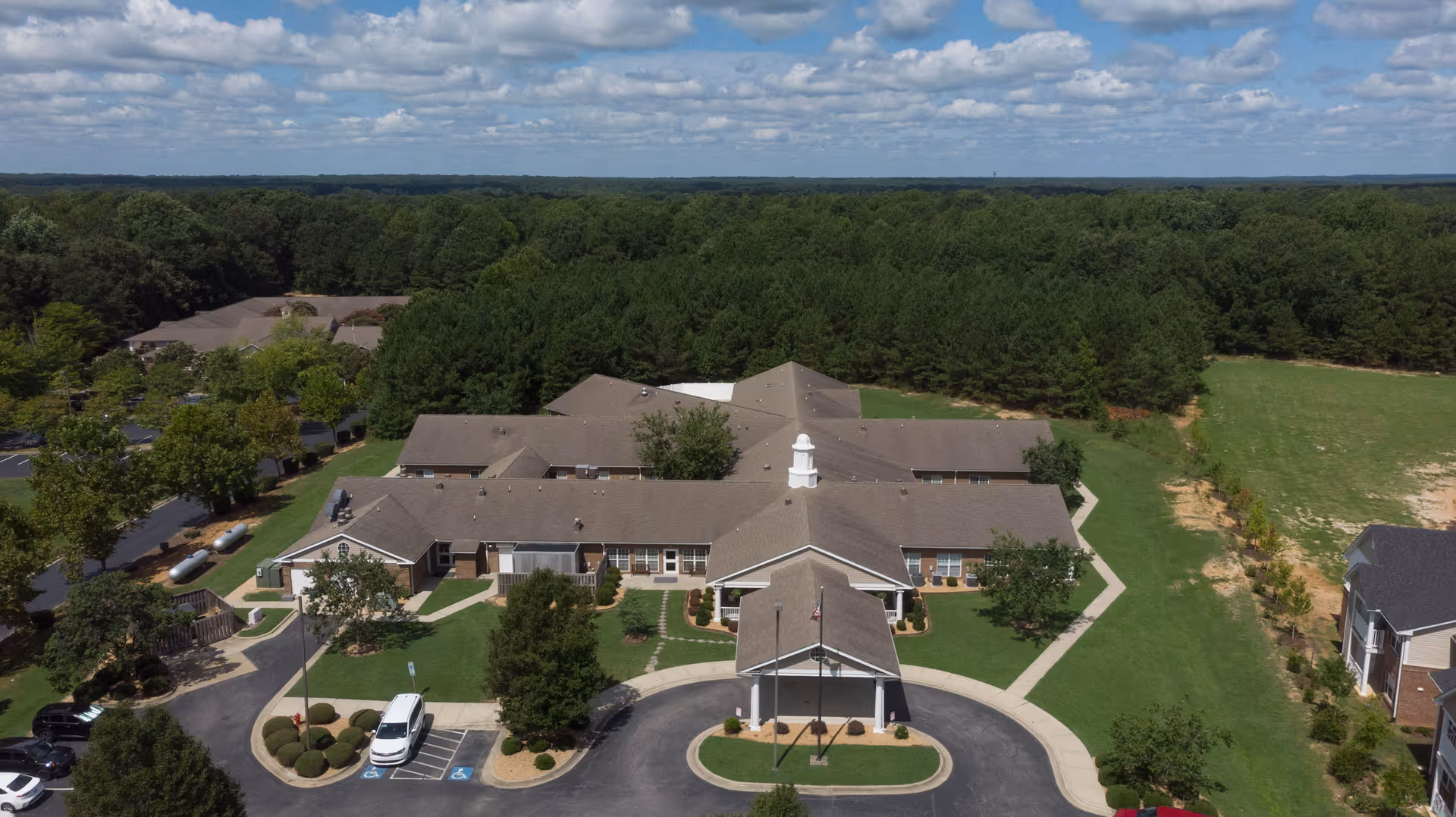 Aerial view of a single-story senior living facility with a circular drive, landscaped grounds, and surrounding trees.