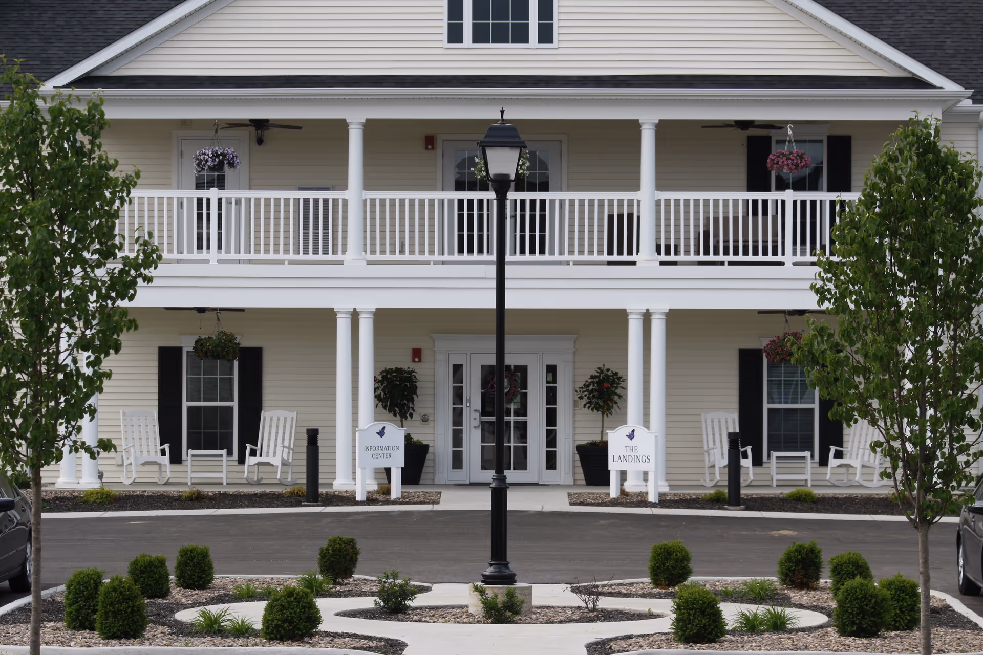 Front exterior view of a senior living community building with a two-story porch, white railings, rocking chairs, hanging flower baskets, and a central lamppost. There are small landscaped bushes and trees in front of the building, with signs reading 'Information Center' and 'The Landings' near the entrance.