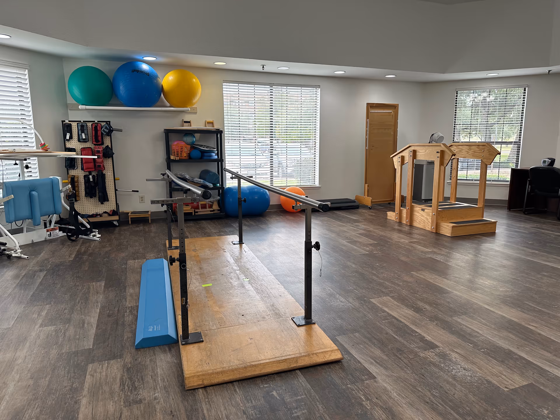 A spacious rehabilitation room with wooden flooring and large windows letting in natural light. The room contains parallel bars for walking exercises, various exercise balls on shelves and the floor, a blue foam wedge, and wooden exercise equipment. There is also a desk with a chair in the corner near the windows.