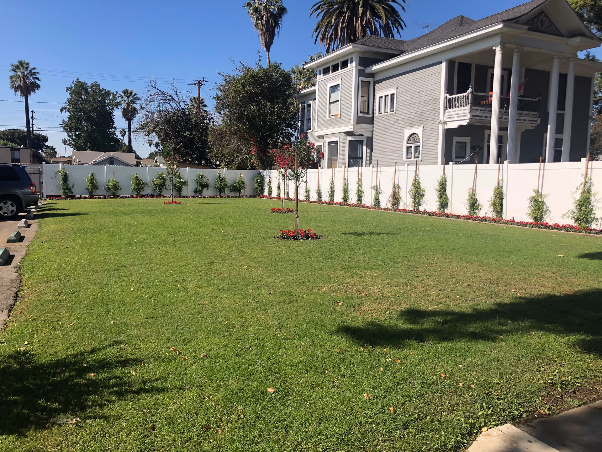 A well-maintained grassy outdoor area with small trees planted in the center and along the white fence. There are palm trees and other greenery in the background, and a large gray two-story house with a balcony is visible on the right side. A parked vehicle is partially visible on the left side.