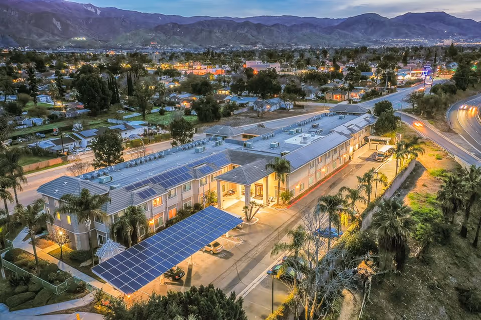 Aerial view of a two-story senior living facility building at dusk with solar panels on the roof and carport, surrounded by trees and residential houses, with mountains in the background and streetlights illuminating the area.