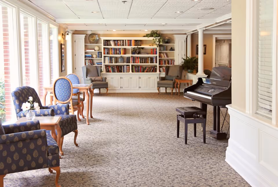 A bright and spacious lounge area with patterned carpet, several upholstered armchairs and small wooden tables near large windows letting in natural light. At the far end, there is a white built-in bookshelf filled with books, flanked by two armchairs. On the right side, there is a black upright piano with a matching bench. The room has a calm and inviting atmosphere.