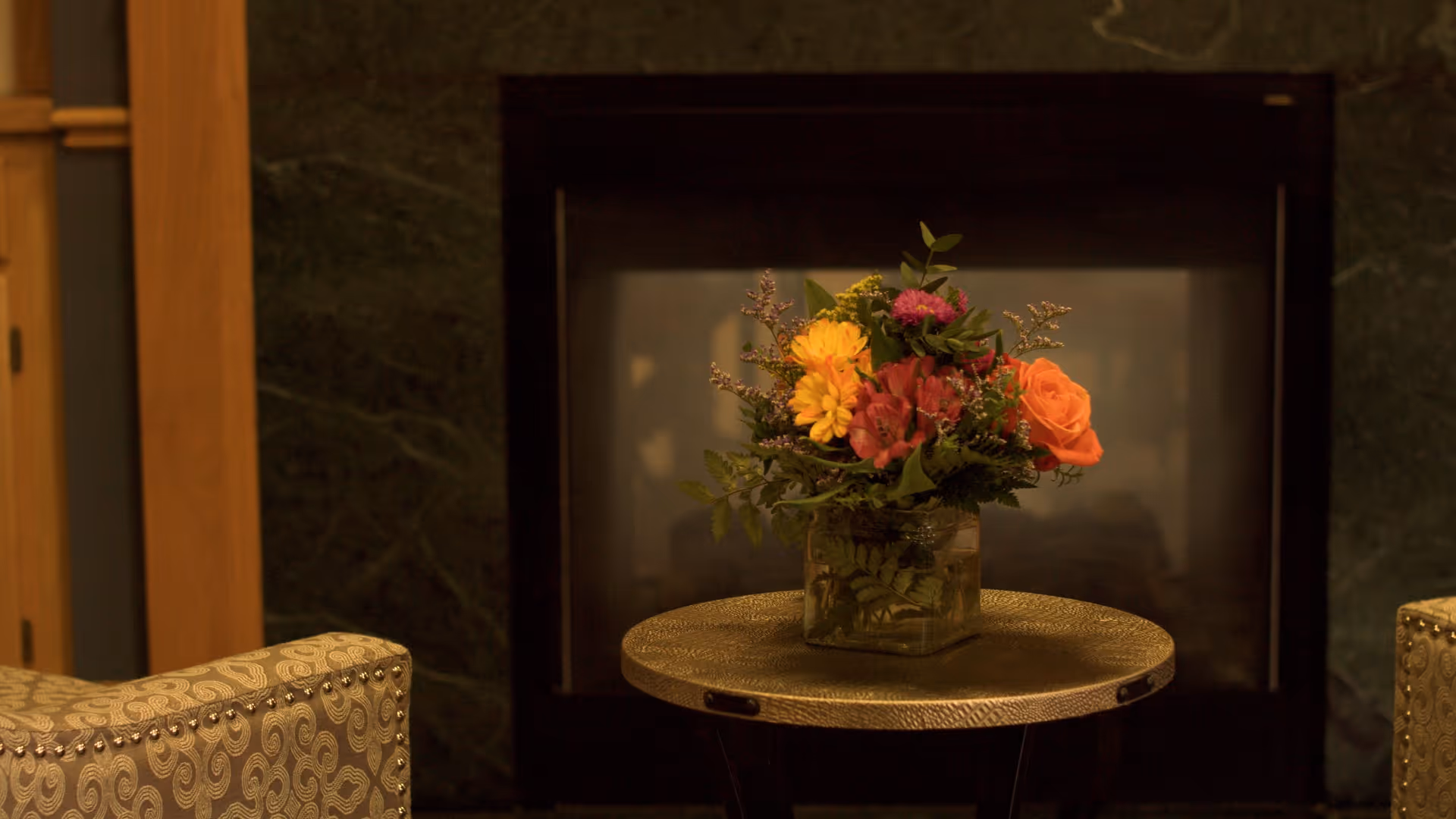 A close-up view of a small round table with a textured gold surface holding a square glass vase filled with a colorful bouquet of flowers, including orange, yellow, and pink blooms, set in front of a dark fireplace with a green marble surround. Part of two upholstered chairs with a patterned fabric and nailhead trim are visible on either side of the table.