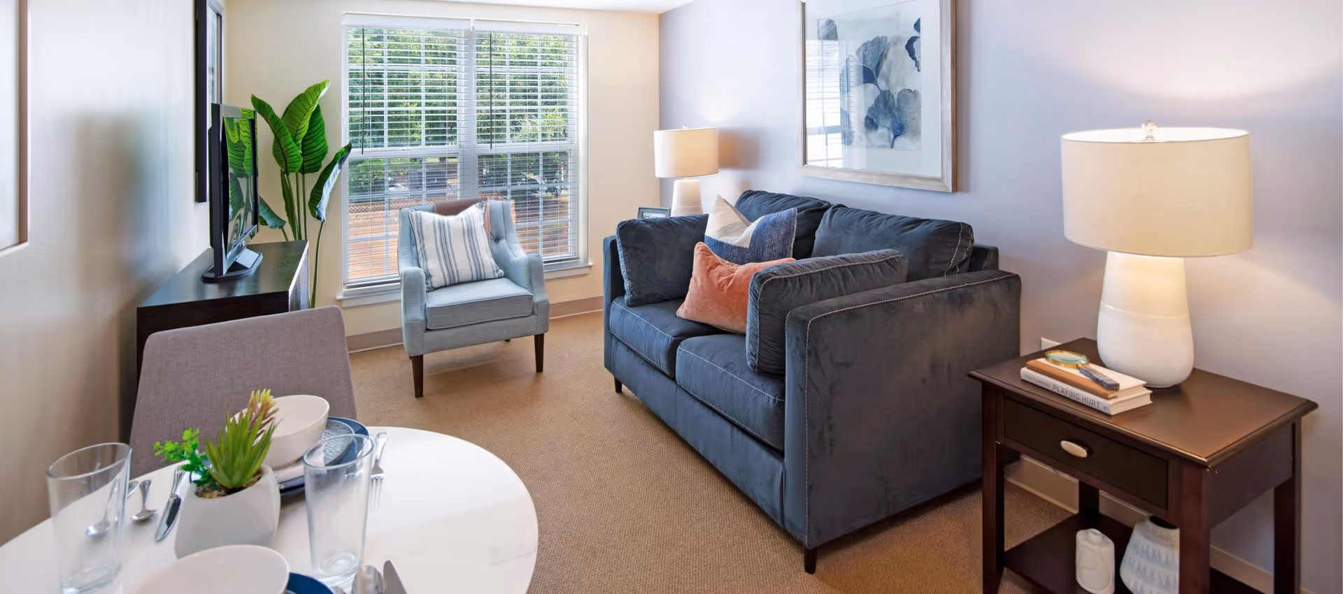 Sunlit living room with a blue sofa and armchair by a window, side table with a lamp, TV on a stand, and a small dining table in the foreground.