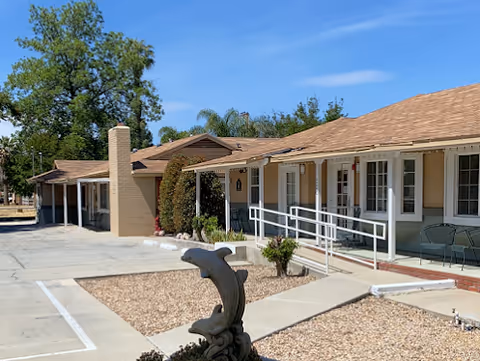 Exterior view of a single-story assisted living facility building with a light brown roof and beige walls. There is a wheelchair-accessible ramp leading to the entrance, some outdoor chairs on the porch, a dolphin statue in the landscaped area, and trees in the background under a clear blue sky.