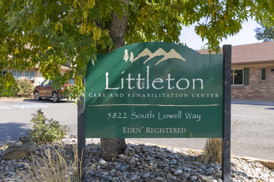 A green sign for Littleton Care and Rehabilitation Center with the address 5822 South Lowell Way, placed in a landscaped area with rocks and plants. A building and parked cars are visible in the background.