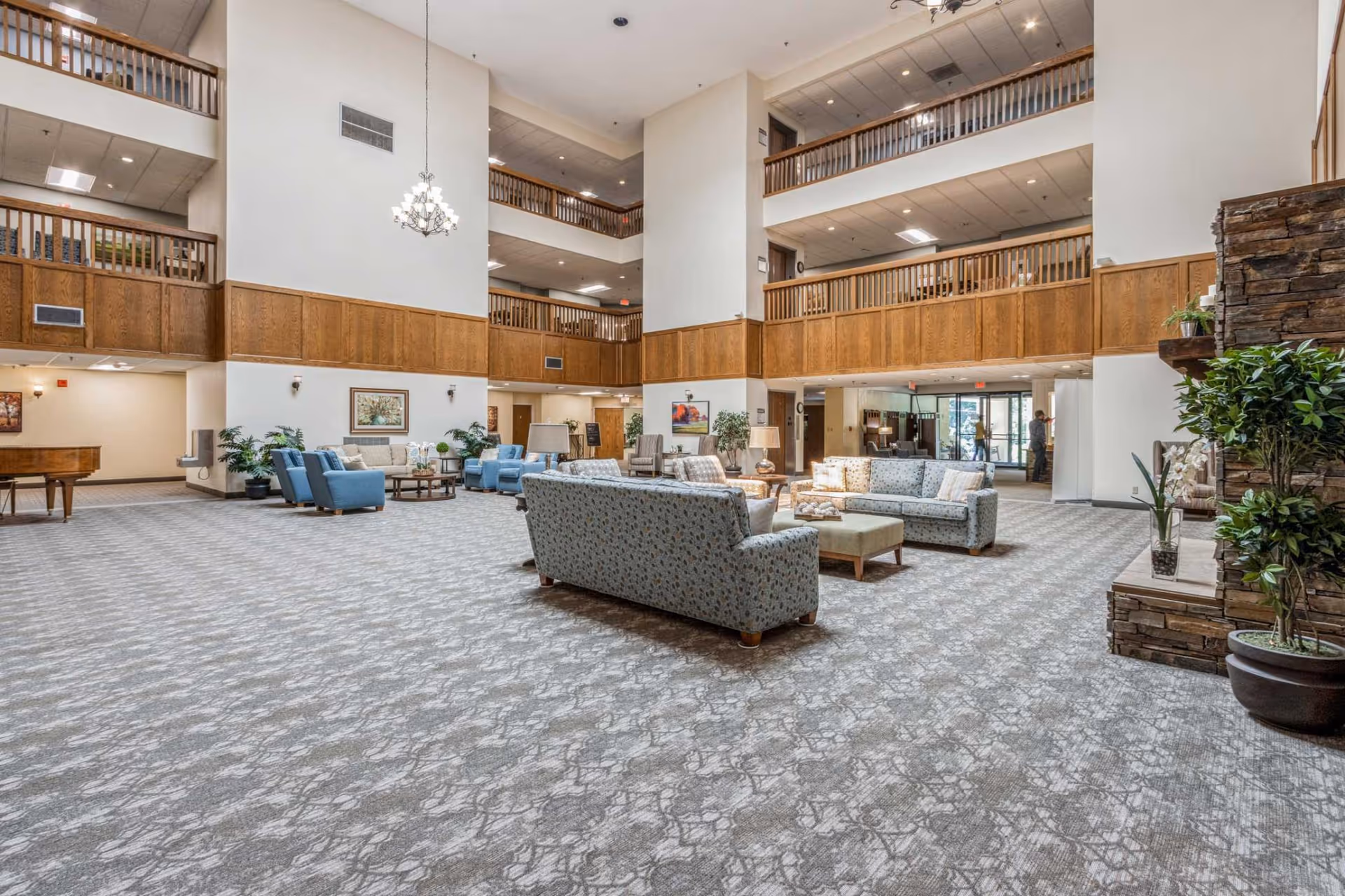 Spacious senior living facility common area with high ceilings, multiple seating arrangements including sofas and armchairs, carpeted floor, wooden railings on the upper floors, and a stone fireplace with plants nearby.
