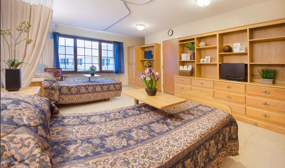 A bright room with two beds covered in patterned bedspreads, a wooden table with a flower vase between the beds, large windows with blue curtains, and a wooden shelving unit with a small TV and decorative items.