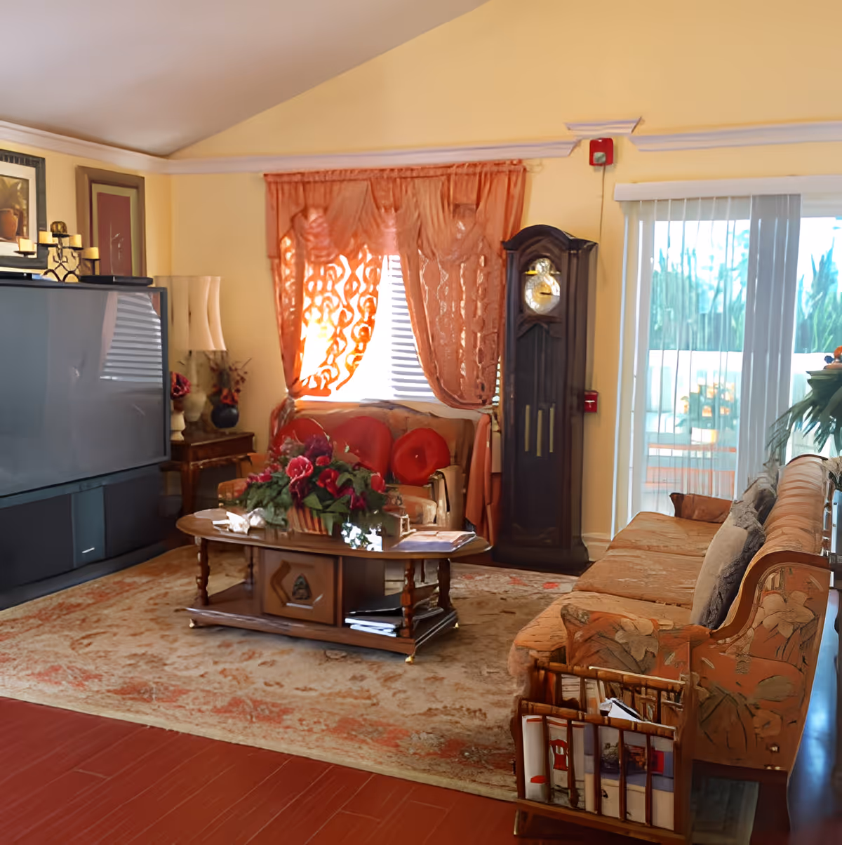 Sunlit living room with patterned sofas, a central coffee table with flowers, a grandfather clock, large TV, and sliding glass doors.