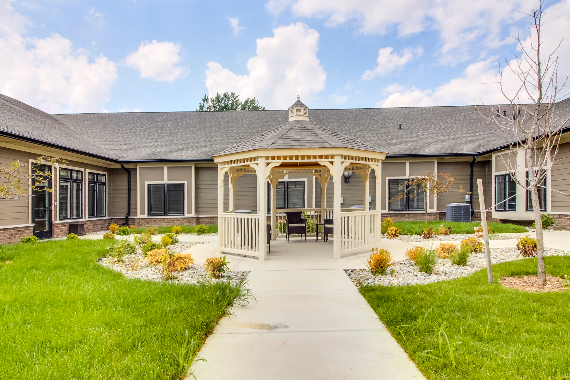 Outdoor courtyard area of Glen Abbey Assisted Living & Memory Care featuring a beige gazebo with chairs and a table inside, surrounded by green grass, small plants, and a concrete walkway leading to the gazebo. The building exterior is visible in the background under a partly cloudy sky.