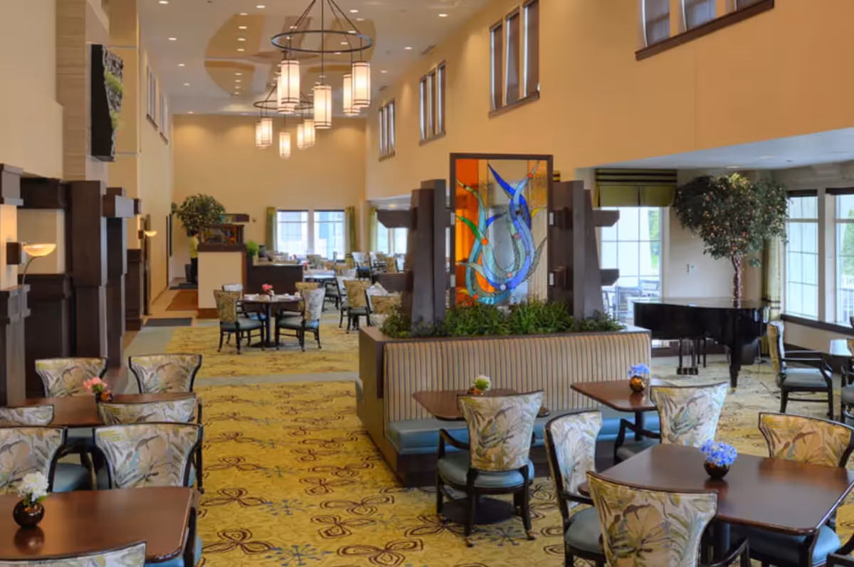 Spacious senior living dining room with multiple tables and patterned chairs, a decorative glass partition, and a grand piano.