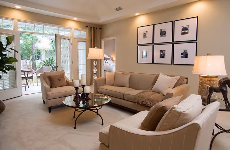 A cozy living room with beige walls and carpet, featuring a beige sofa with cushions and a throw blanket, two beige armchairs, a round glass coffee table with candles, two table lamps, and framed black-and-white photos on the wall. French doors open to an outdoor patio with chairs and greenery visible outside.