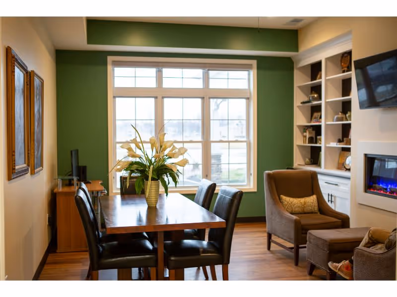 A cozy interior room with a wooden dining table surrounded by four black leather chairs. A vase with white flowers is placed on the table. The room has a large window with multiple panes letting in natural light. To the right, there is a comfortable armchair with a matching ottoman, a built-in white shelving unit with decorative items, and a wall-mounted TV above an electric fireplace. The walls are painted green and beige, and the floor is wooden.