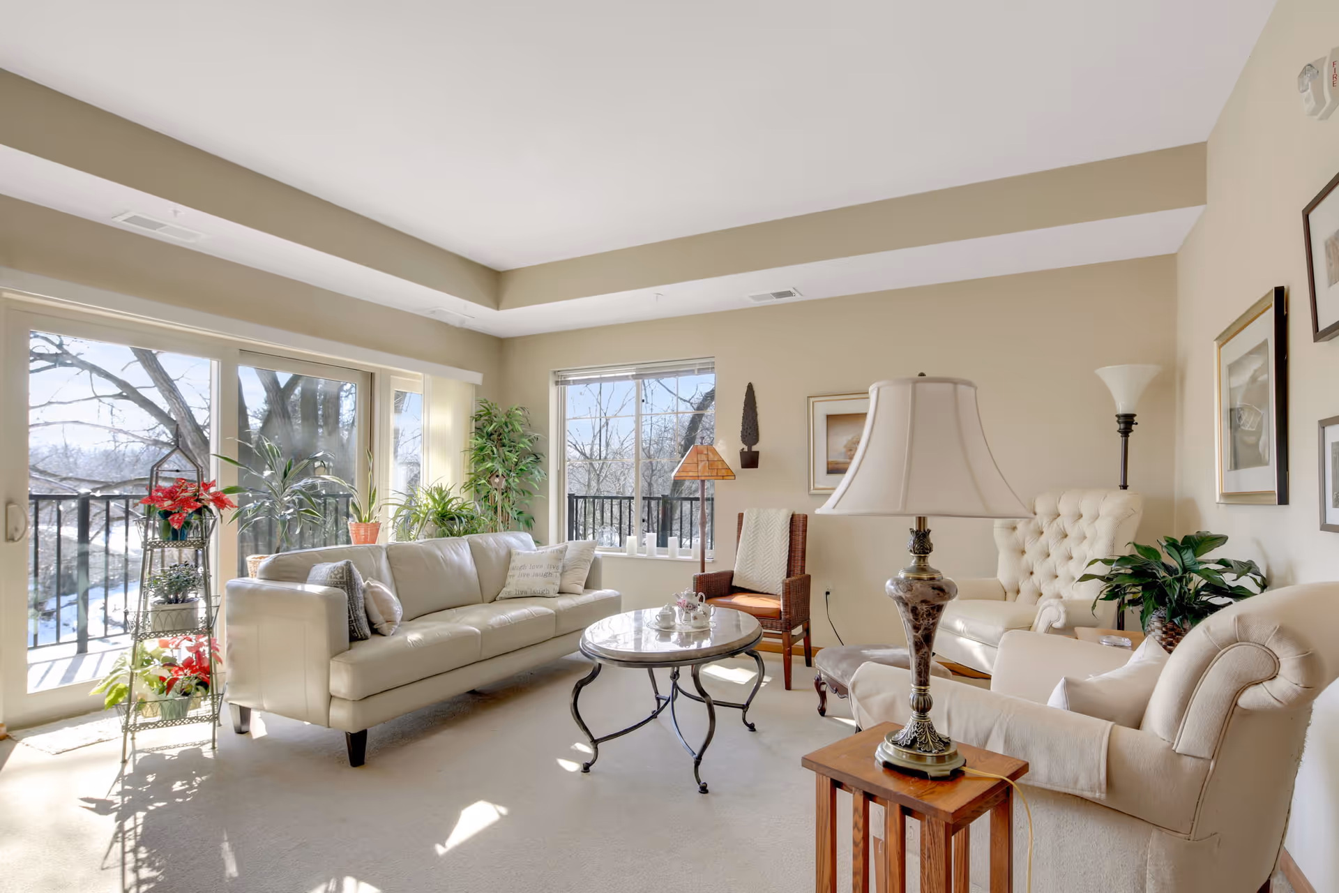 Bright living room with large windows and sliding glass doors letting in natural light. The room features a white leather sofa, a beige armchair, a tufted cream armchair, a wooden side table with a decorative lamp, a round glass coffee table, several potted plants, and framed artwork on the walls.