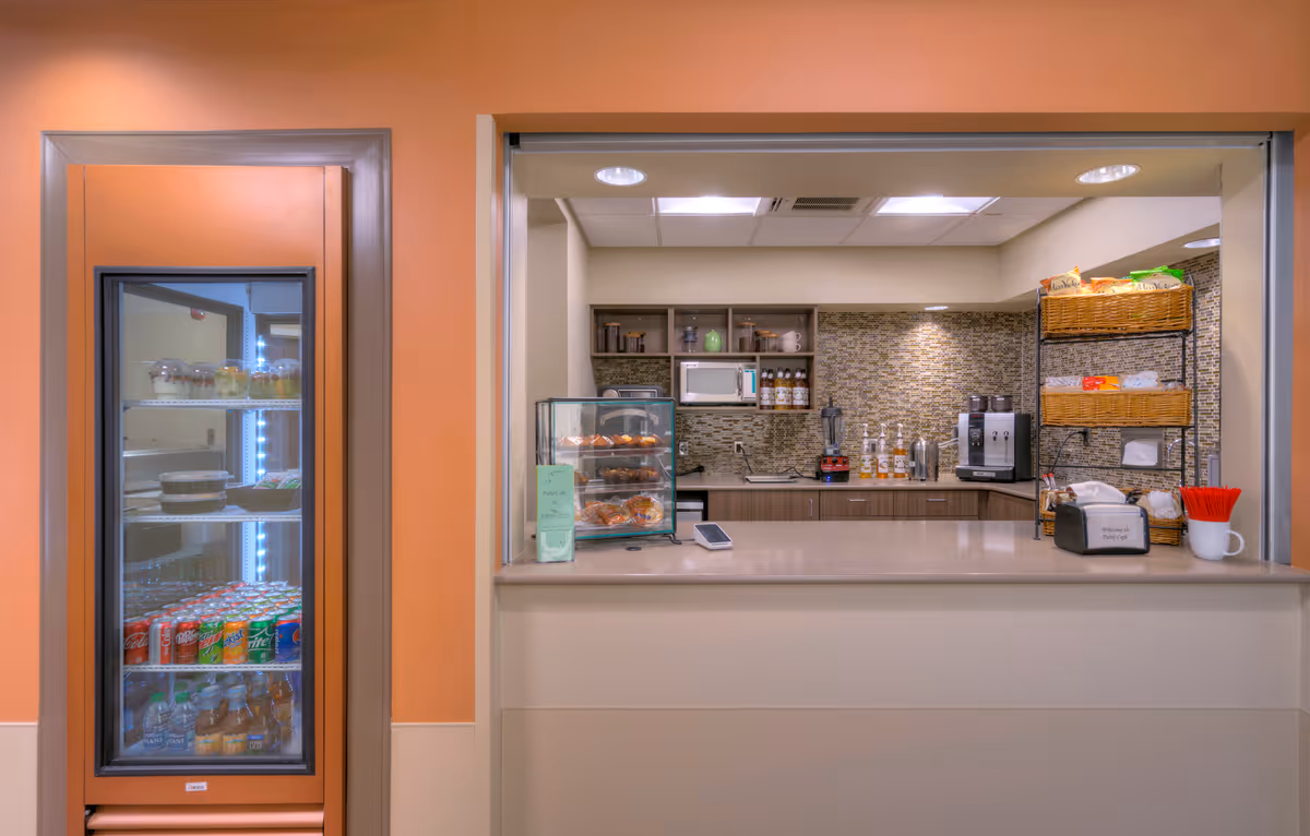 Service counter and snack bar area with a refrigerated display of drinks, pastries, coffee machine, and shelving in a retirement community interior.