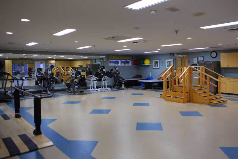 A spacious rehabilitation and exercise room with various fitness equipment including stationary bikes, parallel bars for walking practice, a wooden staircase for physical therapy, and exercise balls on shelves. The room has a light-colored floor with blue square patterns and bright overhead lighting.