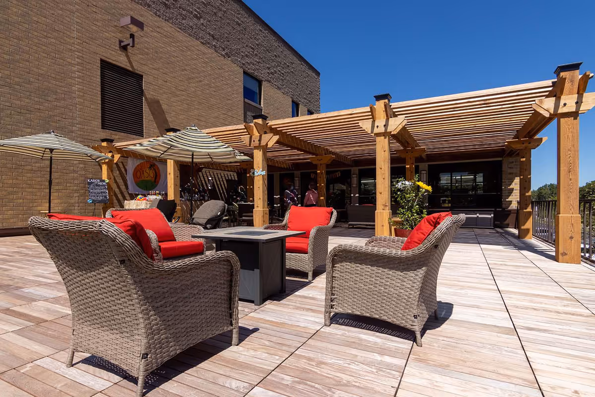 Outdoor patio area with wicker chairs featuring red cushions arranged around a square fire pit table. The patio has a wooden pergola providing partial shade, striped umbrellas, and a brick building wall in the background under a clear blue sky.