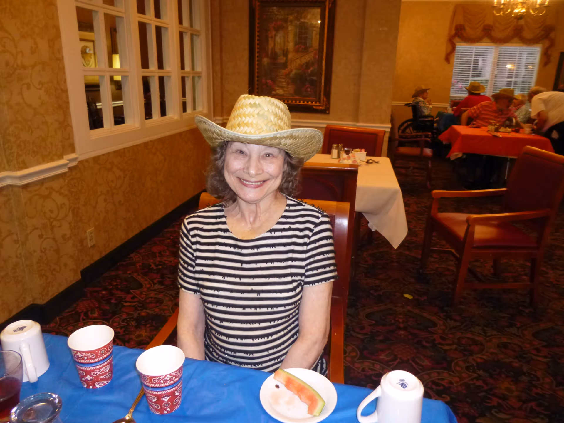 An elderly woman wearing a straw hat and a black and white striped shirt is sitting at a table with a blue tablecloth. On the table are two red patterned paper cups, a white plate with a slice of watermelon, a white mug, and a glass with a dark beverage. In the background, other elderly people are seated at tables with red tablecloths in a warmly decorated dining room with patterned carpet and wallpaper.