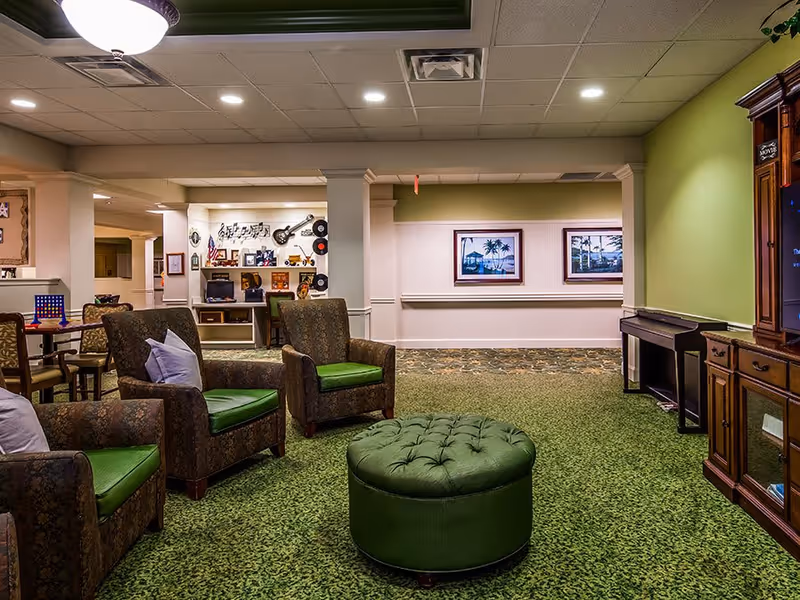 A cozy senior living facility common area with patterned armchairs featuring green cushions arranged around a green tufted ottoman. The room has green carpet, light green walls, and a ceiling light fixture. In the background, there is a small desk area decorated with music-themed wall art, including a guitar and records, and framed pictures on the walls. A wooden entertainment center with a TV is visible on the right side.