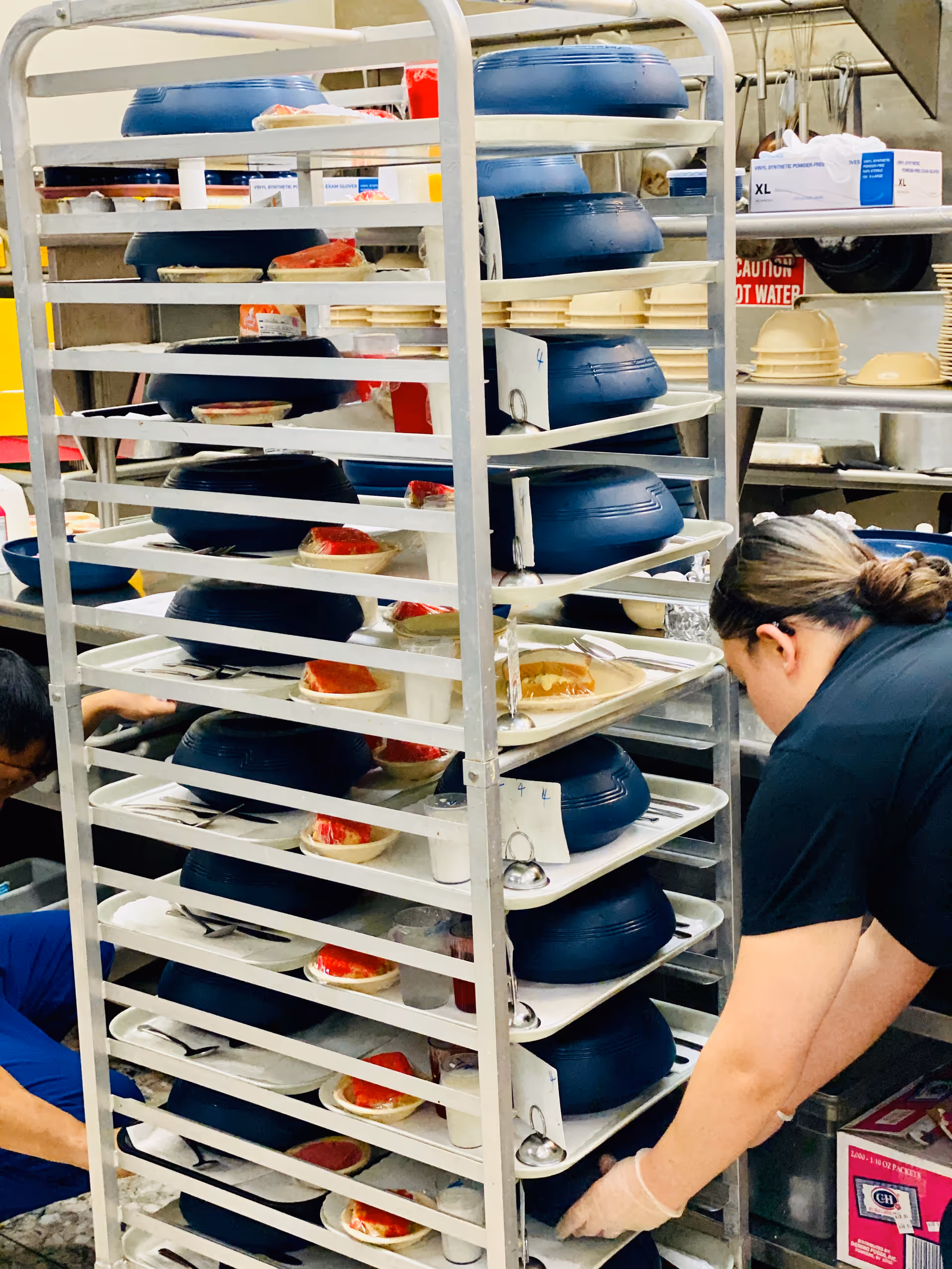 A kitchen scene showing a metal rack filled with trays of covered meals and desserts. Two people are seen working near the rack, organizing or preparing the trays. Various kitchen supplies and utensils are visible in the background.