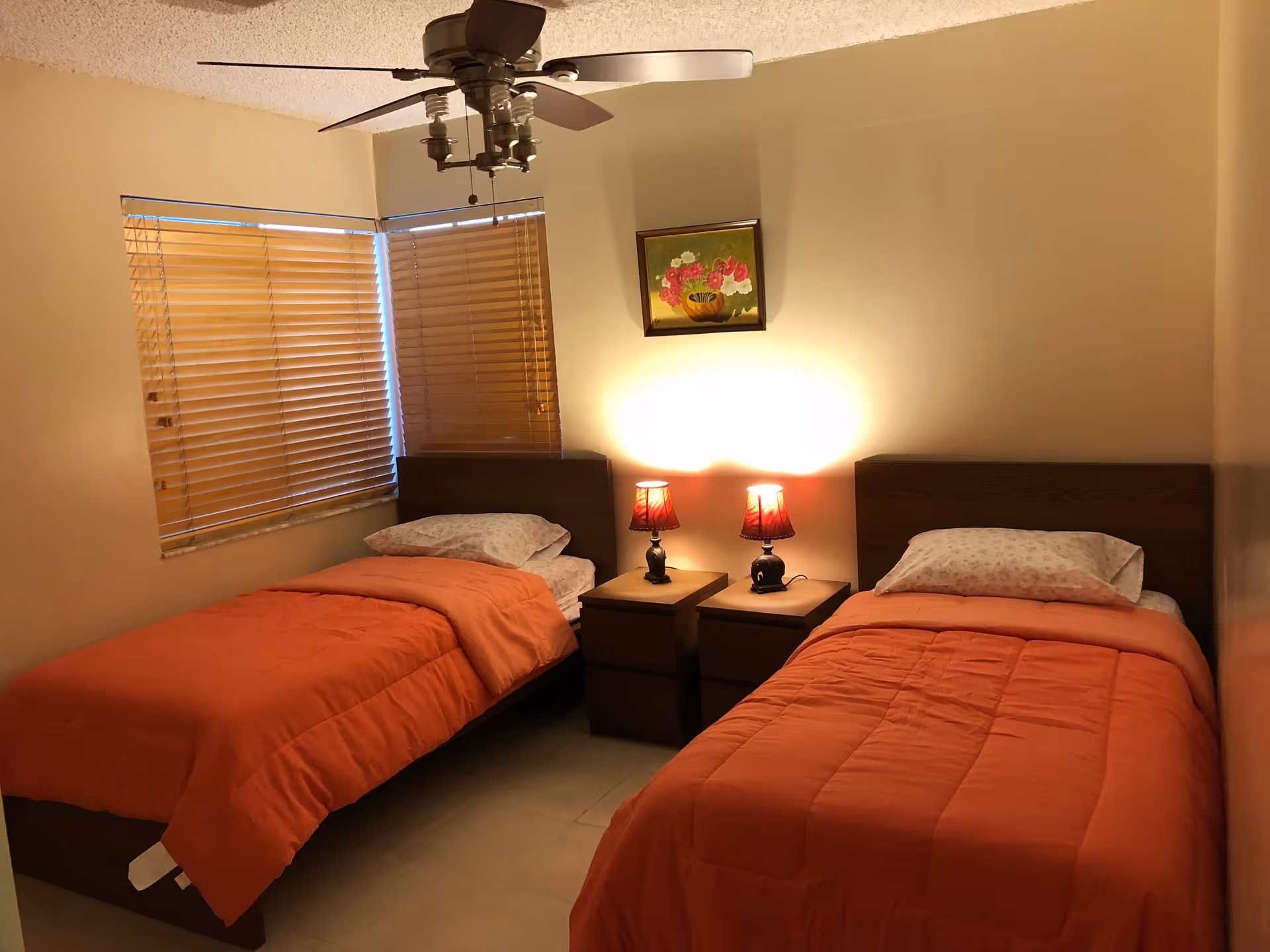 A bedroom with two single beds, each with an orange comforter and a pillow with a floral pillowcase. Between the beds are two wooden nightstands with two small lamps that have red lampshades. Above the nightstands is a framed painting of flowers. The room has beige walls, a ceiling fan with lights, and two windows with wooden blinds.