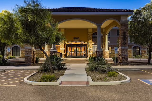Front exterior view of The Mission at Agua Fria Senior Living building entrance at dusk, featuring a covered walkway with stone pillars, trees, and landscaped areas with shrubs and plants.