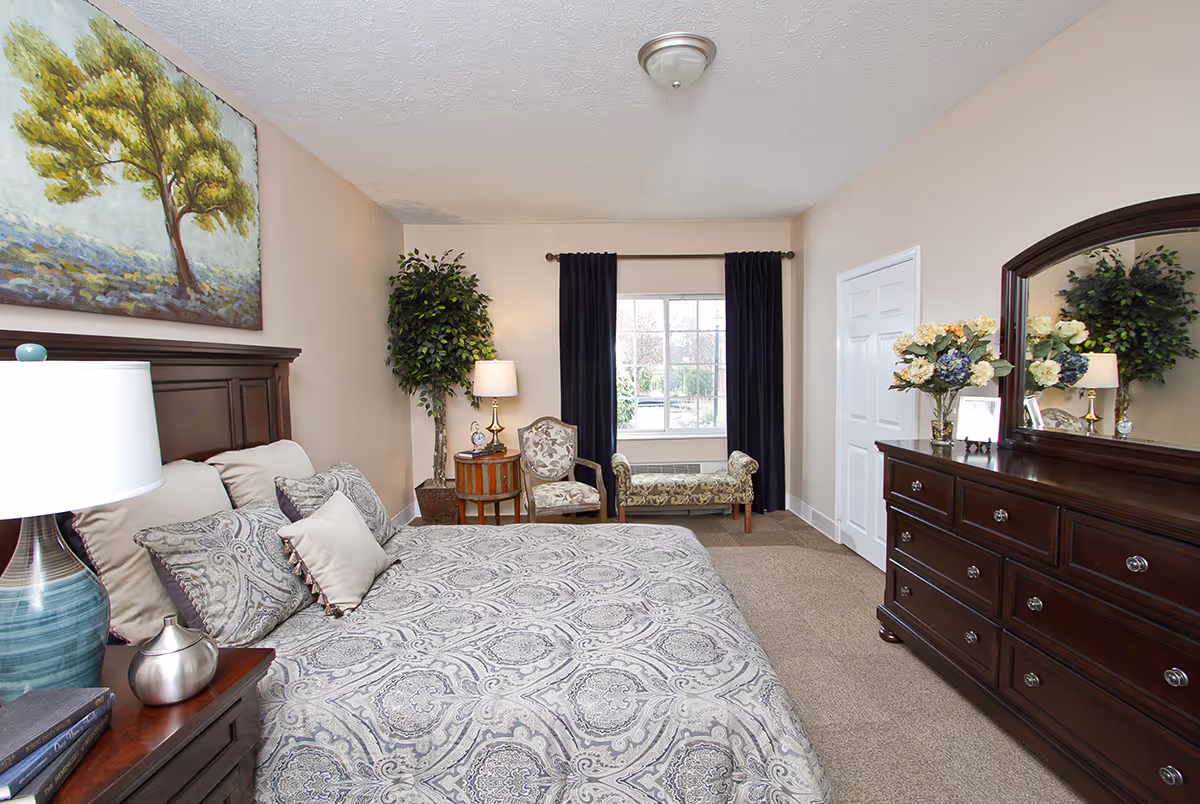 A well-lit bedroom with a large bed covered in patterned bedding, a wooden headboard, and multiple pillows. To the left of the bed is a nightstand with a blue lamp and decorative items. On the far wall, there is a window with dark curtains, a small round table with a lamp, a decorative chair, and a cushioned bench. On the right side of the room is a wooden dresser with a large mirror and a vase of flowers. A large painting of a tree hangs above the bed, and a potted plant is placed near the window.