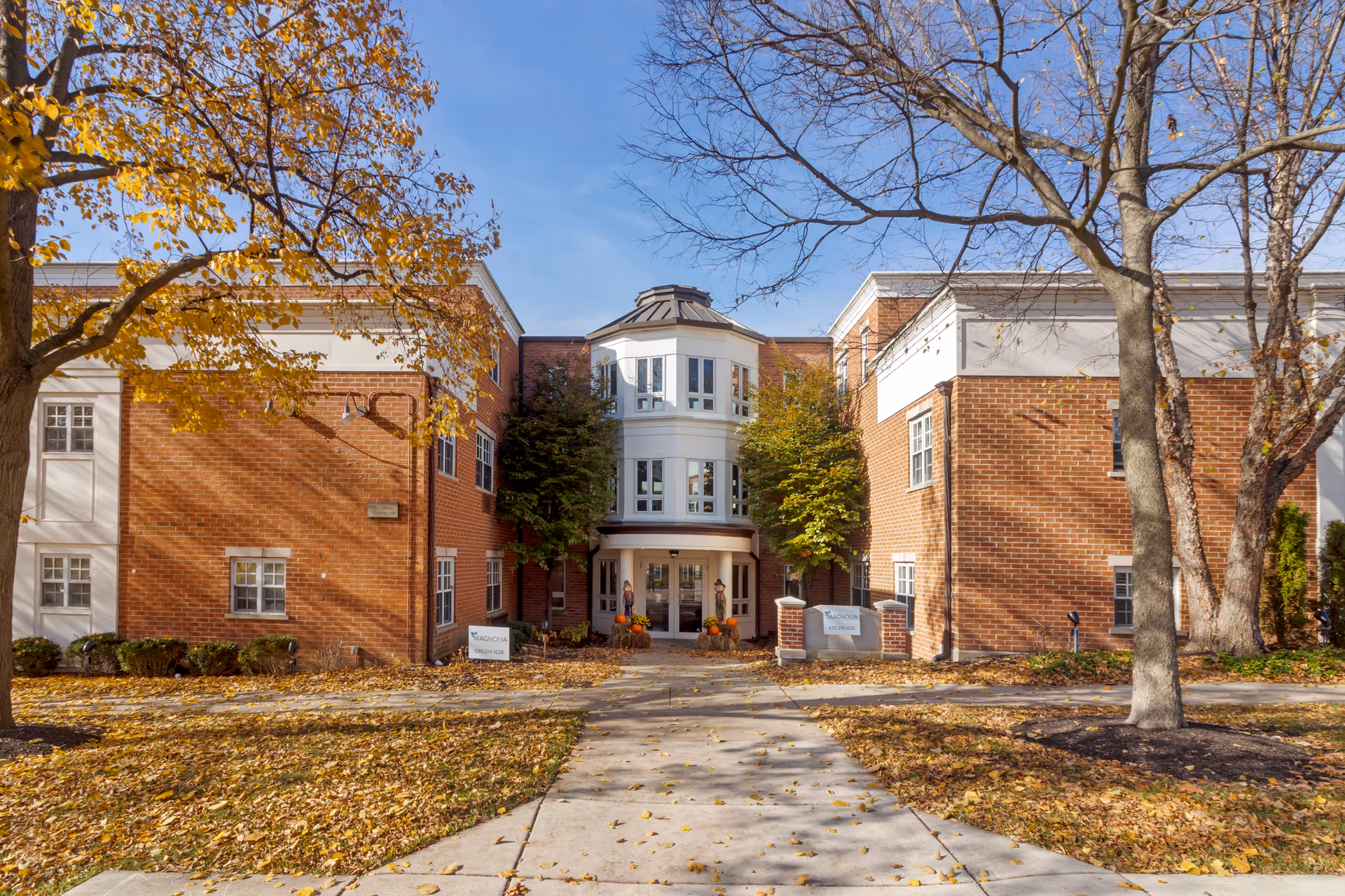 Front exterior view of a brick senior living facility building with a central entrance flanked by two trees with autumn leaves. The building has white trim and multiple windows, with a clear blue sky above.