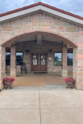 Stone building entrance with an arched brick design, double wooden doors decorated with wreaths, two windows on either side, a rocking chair on the left, and two flower pots with pink flowers on either side of the entrance.
